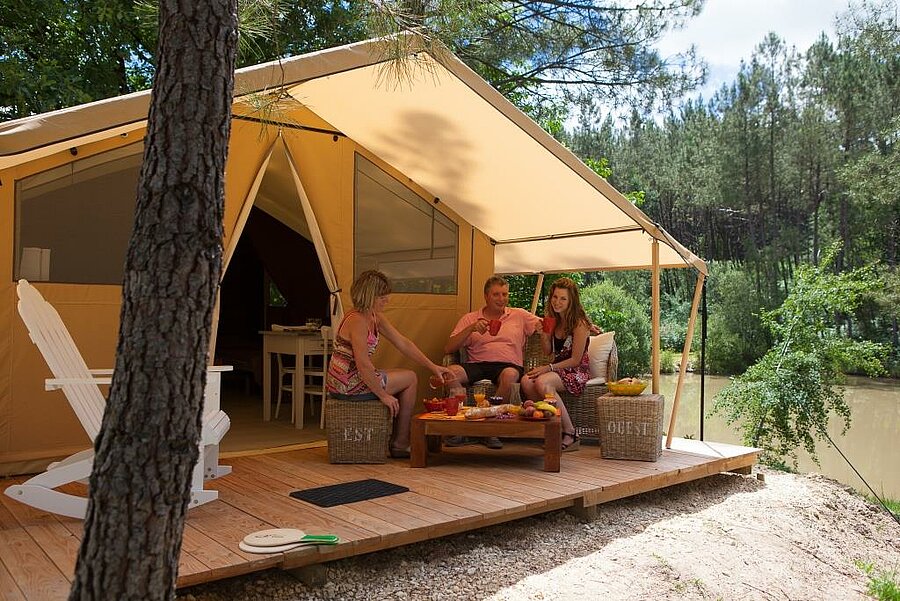 Family enjoying a wooden terrace in front of a lodge tent at a campsite in Nouvelle-Aquitaine, by a lake and surrounded by pine trees.